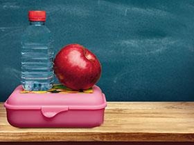 Butelka wody i jabłko stojące na pojemniku na drugie śniadanie, w tle tablica szkolna