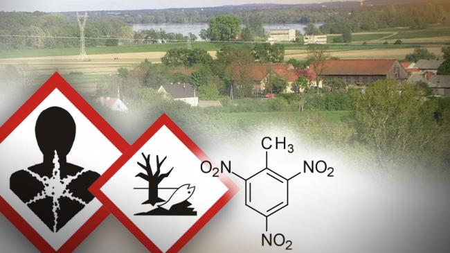 Znaki Produkt poważnie zagrażający zdrowiu i Produkt niebezpieczny dla środowiska obok wzór chemiczny trotylu, w tle widok na dzielnicę Łęgnowo