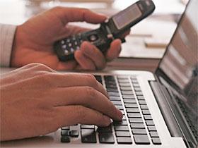 Dłonie nad klawiaturą laptopa, w jednej z dłoni telefon komórkowy