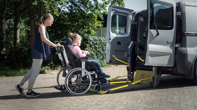 Ilustracja: Dziewczynka na wózku inwalidzkim jest wprowadzana do na rampę samochodu przystosowanego do przewozu osób na wózkach