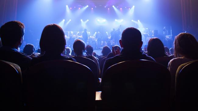 Publiczność obserwująca oświetlaną scenę, na której muzycy przygotowują się do koncertu
