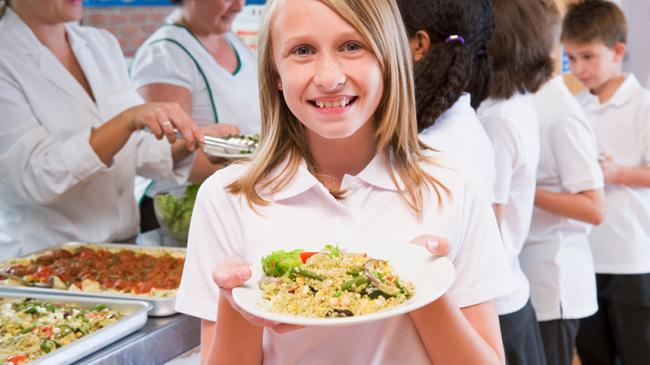 Dziewczynka na szkolnej stołówce pokazuje zawartość swojego talerza, za nią kolejka innych dzieci odbierających posiłek