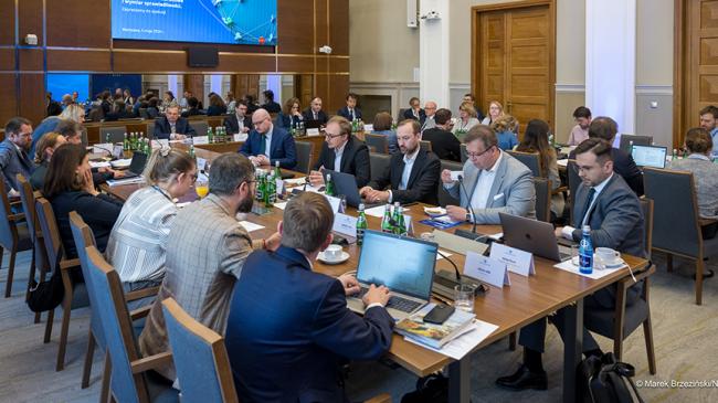 Uczestnicy panelu siedzący przy stołach, nad nimi w tle ekran wyświetlający przedmiot dyskusji