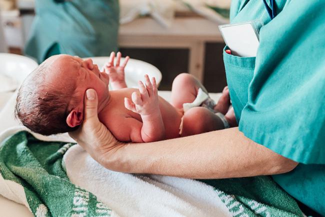 Noworodek tuż po porodzie jest delikatnie trzymany na rękach przez pracownika medycznego w zielonym stroju. Dziecko leży na ręczniku w sali porodowej, a w tle widoczne jest wyposażenie szpitalne