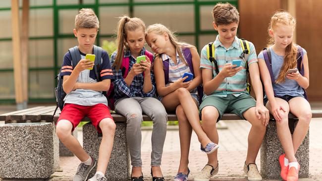 Pięcioro dzieci z plecakami siedzi na betonowej ławce przed budynkiem i patrzy w ekrany smartfonów trzymanych w dłoniach