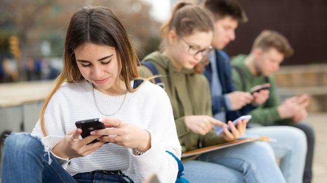 Grupa młodych osób siedzących na zewnątrz i korzystających ze smartfonów.