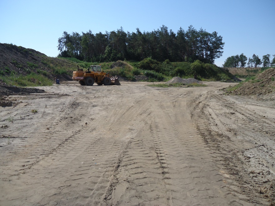 Wyrobisko Lewkówka I&nbsp;- spychacz na&nbsp;rozjeżdżonej ziemi, w tle porośnięte trawą hałdy